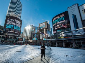 A pedestrian walks through Dundas Square in Toronto with digital signage stating "Stay Home Stop The Spread Save Lives" and "Restarting Our Economy Starts By Staying Home,” during the COVID-19 pandemic, in 2021.