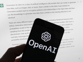 The OpenAI logo is seen on a mobile phone in front of a computer screen displaying output from ChatGPT.