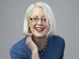 A portrait of Laura Feltz, the subject of this article and who lives with low vision from glaucoma. A woman with white-blond hair cut to a bob smiles for the camera. She is wearing black glasses and a blue shirt with a gold necklace.