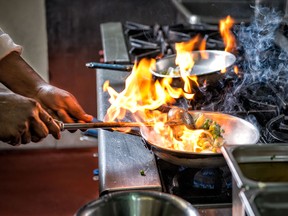 A chef cooking food in frying pans over an open flame in a restaurant kitchen