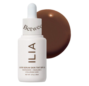 Upright dropper bottle of ILIA Super Serum Skin Tint SPF 40 Foundation with a large drop of sunscreen in background.