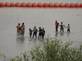Migrants who crossed the Rio Grande from Mexico walk past large buoys being deployed as a border barrier on the river in Eagle Pass, Texas, July 12.