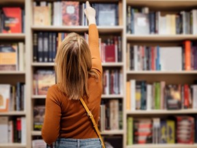 Girl with loose long blondie hair standing and holding an open book between book shelves in the library, reading and looking into the book.