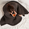 An overhead shot of a woman sitting, completely wrapped in a dark grey-brown hand knit weighted blanket, holding a cup of tea.