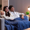A man and a woman cuddled up on a couch, under a dark blue weighted blanket presumably watching TV.