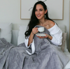 A woman sitting up in bed holding a cup of tea, with a grey weighted blanket draped over her lap.