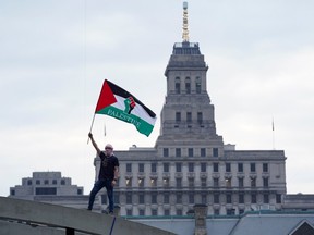 A protester waves a Palestine flag from on top of an arch at Nathan Phillips Square