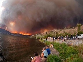 People at the edge of a lake watch a wildfire in the distance.