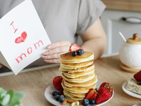 A stack of pancakes with blueberries and strawberries on a table in front of a person reading a card that says 'I heart mom' on the front.