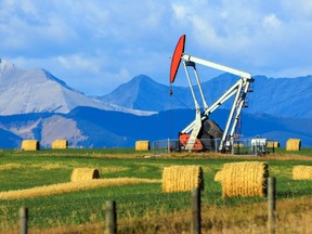 An oil and gas industry pumpjack drill rig in a field with hay bales in the foreground and mountains in the background.