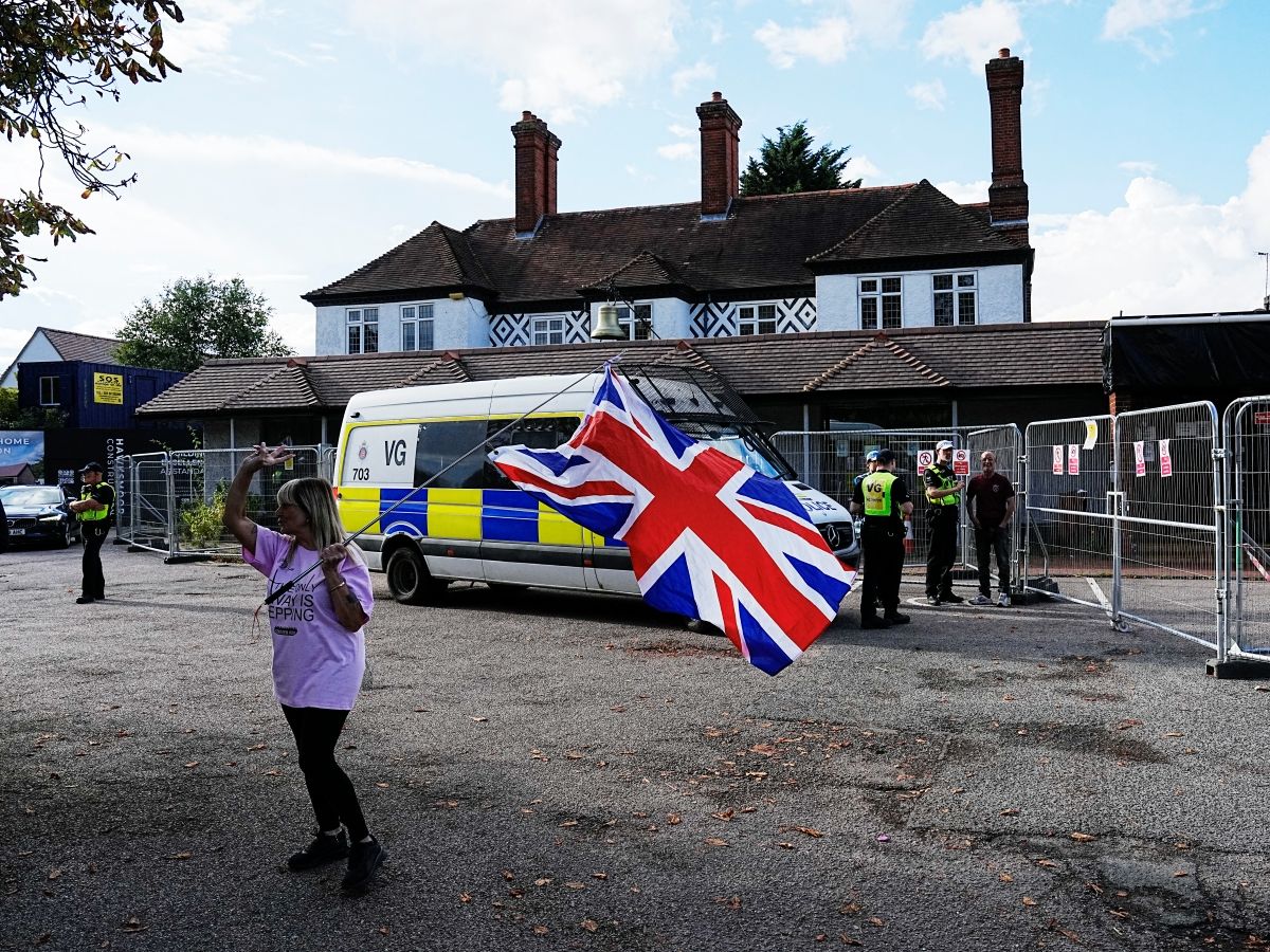 Police officers sercure the area as a protester dubbed one of the 'Pink Ladies' hold a union flag outside the Bell Hotel in Epping on August 29, 2025, after the appeals court overturned a decision temporarily blocking the use of the hotel to house asylum seekers.