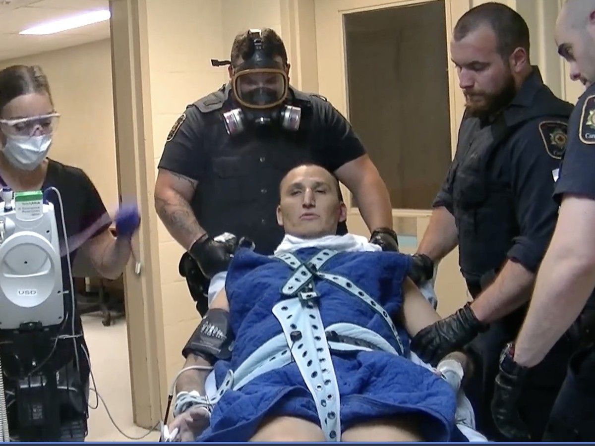 A man is tied down to a stretcher by guards in a prison