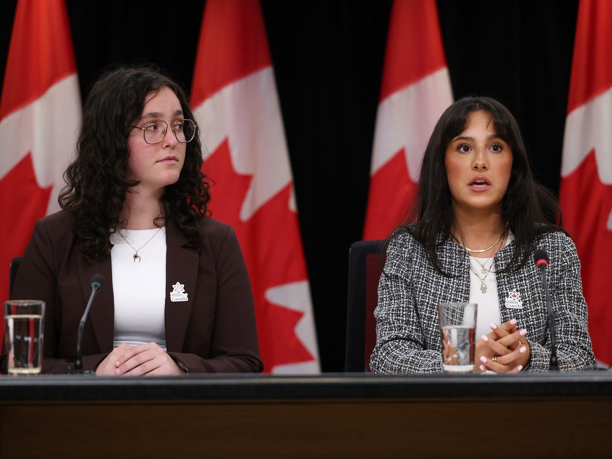 Two young women speak from behind a desk at a press conference with Canadian flags in the background.