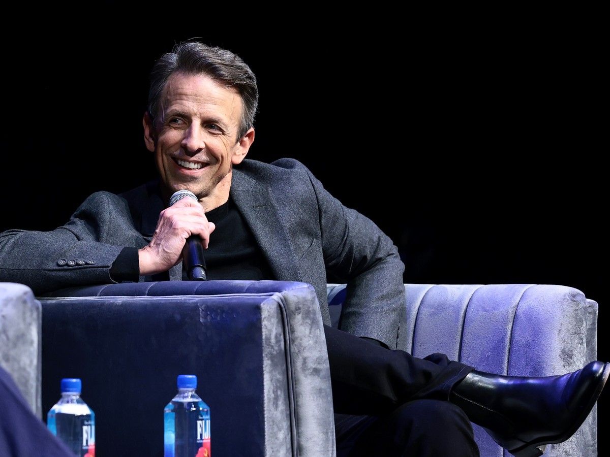 Seth Meyers sitting on a couch and holding a microphone.