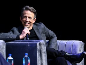 Seth Meyers sitting on a couch and holding a microphone.