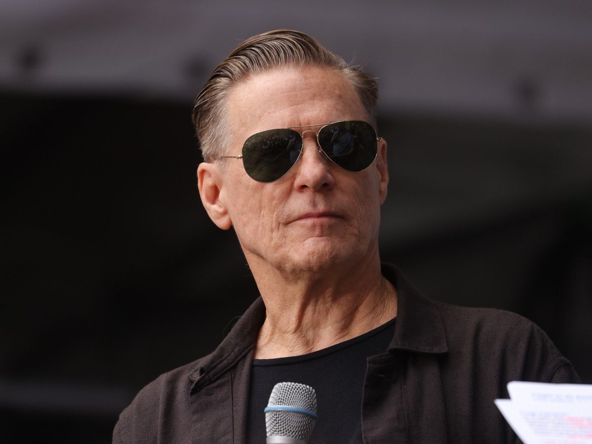 Bryan Adams pictured wearing sunglasses and standing behind a microphone.