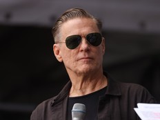 Bryan Adams pictured wearing sunglasses and standing behind a microphone.