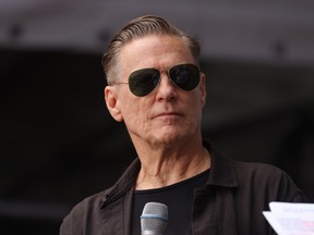 Bryan Adams pictured wearing sunglasses and standing behind a microphone.
