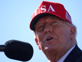 A close up of Donald Trump wearing a USA hat and speaking into a microphone in front of a blue sky.