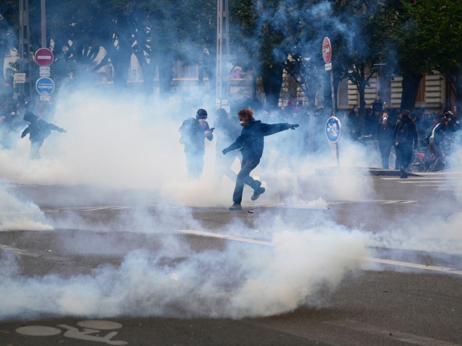 A demonstrator attempts to kick away a fuming tear gas pellet amid a cloud a of tear gas during a demonstration in favor of purchasing power, the protection of public services, and fiscal and social justice, in Lyon, central France on October 24, 2026 as part of a nationwide call for mobilisation bringing together former members of the Yellow Vest movement, labor unions, high school and university students