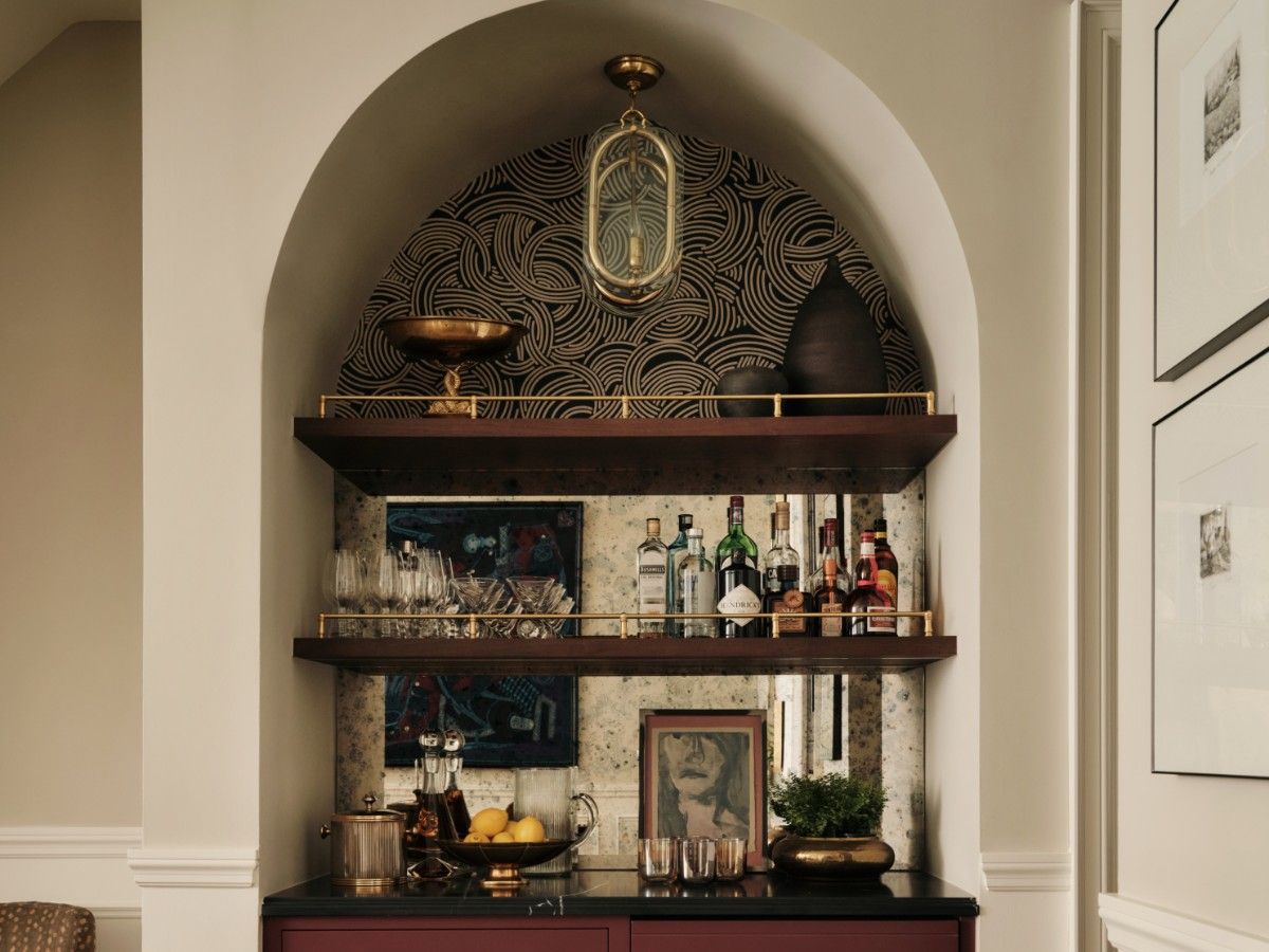 The bar niche is a spirited composition of Schumacher’s turquoise Eastern Eden wallpaper, wine-toned Benjamin Moore New London Burgundy cabinetry, wood shelving and a black work surface, with an antique mirror and vintage gold-and-glass light fixture.