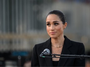 Megan Markle wearing a black jacket and standing behind a microphone.