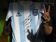 People flash the V sign next to a replica of Argentina's football team jersey used during the final of the FIFA World Cup Mexico '86, handwritten and signed by Argentine former football star Diego Maradona, at a community eatery during the lockdown imposed by the government against the spread of the new coronavirus, COVID-19, in Jose C. Paz, Buenos Aires, Argentina, on May 8, 2020. - Maradona's signed jersey will be raffled among those who donate food to feed people at a working class neighbourhood called Rene Favaloro, after the Argentine cardiac surgeon. (Photo by JUAN MABROMATA / AFP) (Photo by JUAN MABROMATA/AFP via Getty Images)