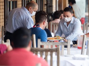 A waiter wearing a mask and gloves delivers food to a table to customers seated at an outdoor patio.