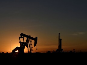 A pump jack operates in front of a drilling rig at sunset in an oil field.