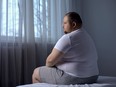 Depressed fat man sitting on bed at home, worried about overweight, insecurities
