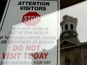 A sign on a downtown business warns people with COVID-19 symptoms to keep away while the city hall clock tower is reflected in its window on Monday afternoon. (RONALD ZAJAC/The Recorder and Times)