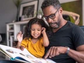 A loving father sits on the couch at home and reads a storybook to his preschool age daughter. The child is sitting on her father's lap and is smiling while looking at the book.