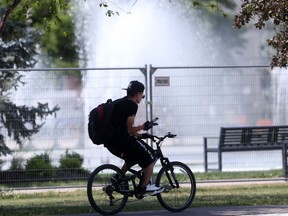 A person ride a bike past a fountain that is behind a fence in Winnipeg on Saturday, July 3, 2021.