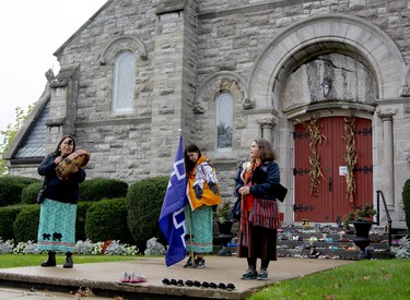 Shannon Olivier, left, Kiki Olivier and Clarice Gervais stand in front of St. John the Evangelist Catholic Church singing and playing the drum during the First Peoples Performing Arts Festival Water Walk. The little shoes memorial that was once at the doors of Gananoque Town Hall was placed at the church for the Water Walk. (JESSICA MUNRO/Local Journalism Initiative)