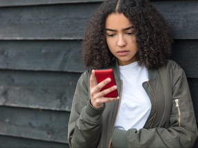 Outdoor portrait of beautiful sad depressed mixed race African American girl teenager female young woman texting on red cell phone wearing green bomber jacket
