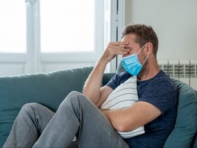 Sad man with protective face mask at home living room couch feeling tired and worried suffering depression amid coronavirus lockdown and social distancing. Mental Health and isolation concept.