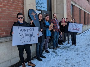 A group of people stand in front of a brick building on a snow covered side walk. They are holding signs that say "Support National GLI!" "I was hungry," "I was cold," and "Put poverty in the grave." One person is hold a cardboard coffin with a cross on it.