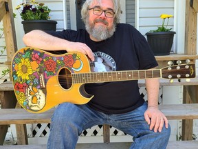 A white man holds a decorated guitar while sitting on porch steps.