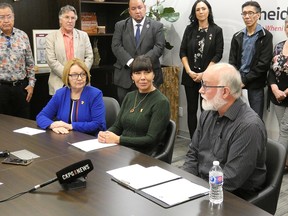 Three people sitting at a conference table with a crowd standing behind.