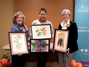 Artist Celeste Noah, centre, of Eelunaapèewii Lahkèewiit, Delaware Nation, holds Berries and Altar, two of her four watercolours now hanging at Chatham-Kent Health Alliance's Chatham site. CKHA board chair Deb Crawford, left, holds the painting Wild Rice, while president and chief executive Lori Marshall holds Indian Corn. (Supplied)