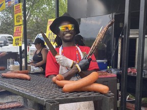 A man in a floppy black hat and red T-shirt stands behind a grill, arms crossed at his chest, while holding a large knife in one hand and a rack of ribs in the other.