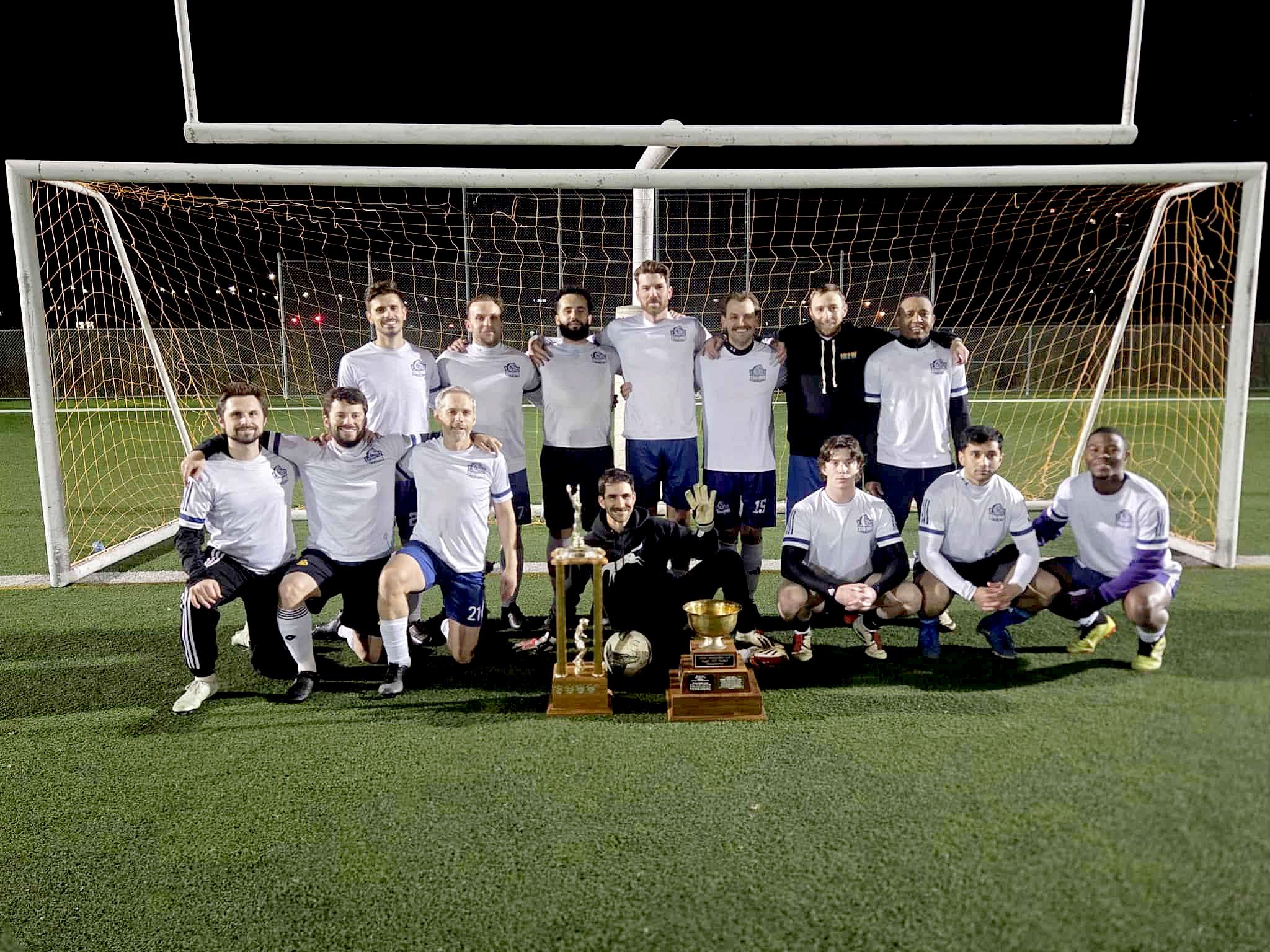A soccer team poses for a photo in front of the net with trophies