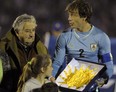 FILE - In this June 4, 2014, file photo, Uruguay's President Jose Mujica, left, gives Uruguay's captain Diego Lugano the national flag before an international friendly soccer match against Slovenia in Montevideo, Uruguay. Uruguay's famously casual President is tossing some earthy expletives at FIFA as he welcomed Uruguay's team back from the World Cup oon Sunday June 29, over its four-month ban of Luis Suarez. Uruguayans overwhelmingly criticized FIFA's decision to ban Suarez from nine national team games and four months for biting an Italian defender. (AP Photo/Matilde Campodonico, File)