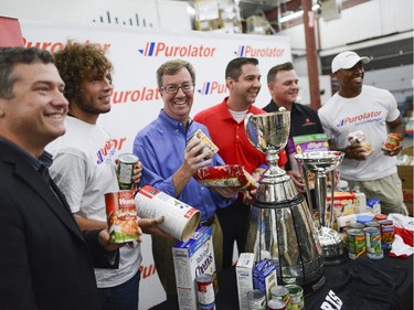 (L-R) Ottawa City Councillor Tim Tierney, Fury FC forwards, Tom Heinemann, Mayor Jim Watson, Manager of corporate sponsorship from Purolator, Brian Collins, Executive director at the Ottawa Food Bank, Michael Maidment, and The Ottawa  Redblacks QB, Henry Burris smiles for the camera at the Ottawa Food Bank for Purolator Tackle Hunger weekend on Thursday, Aug. 14, 2014.  Fans attending the Redblacks and Fury FC games in Aug. 15 and 17 are encouraged to bring non-perishable food items or cash donations to the volunteers stationed at the stadium gates. Fans will have the opportunity to have a photo taken with the Grey Cup and North American Soccer League (NASL) Soccer Bowl trophy.