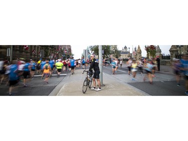 A man stands on the sidewalk with his bike near Parliament Hill as racers make there way past him in both directions on Wellington Street.