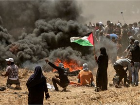 TOPSHOT - A Palestinian woman holding her national flag looks at clashes with Israeli forces near the border between the Gaza strip and Israel east of Gaza City on May 14, 2018, as Palestinians protest over the inauguration of the US embassy following its controversial move to Jerusalem. Dozens of Palestinians were killed by Israeli fire on May 14 as tens of thousands protested and clashes erupted along the Gaza border against the US transfer of its embassy to Jerusalem, after months of global outcry, Palestinian anger and exuberant praise from Israelis over President Donald Trump's decision tossing aside decades of precedent.