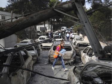 A woman carries bottles of water as people stand amid the charred remains of burned-out cars in Mati east of Athens, Tuesday, July 24, 2018. Twin wildfires raging through popular seaside areas near the Greek capital have torched homes, cars and forests and killed at least 49 people, authorities said Tuesday, raising the death toll after rescue crews reported finding the bodies of more than 20 people huddled together near a beach.