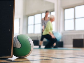 Shot of medicine ball lying on gym floor with senior woman and her personal trainer exercising in background.
