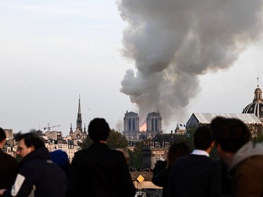 People watch the landmark Notre-Dame Cathedral burning in central Paris on April 15, 2019. - A huge fire swept through the roof of the famed Notre-Dame Cathedral in central Paris on April 15, 2019, sending flames and huge clouds of grey smoke billowing into the sky. The flames and smoke plumed from the spire and roof of the gothic cathedral, visited by millions of people a year. A spokesman for the cathedral told AFP that the wooden structure supporting the roof was being gutted by the blaze.