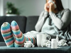 Sick woman with flu, cold, fever and cough sitting on couch at home. Ill person blowing nose and sneezing with tissue and handkerchief. Woolen socks and medicine. Infection in winter. Resting on sofa. Getty Images